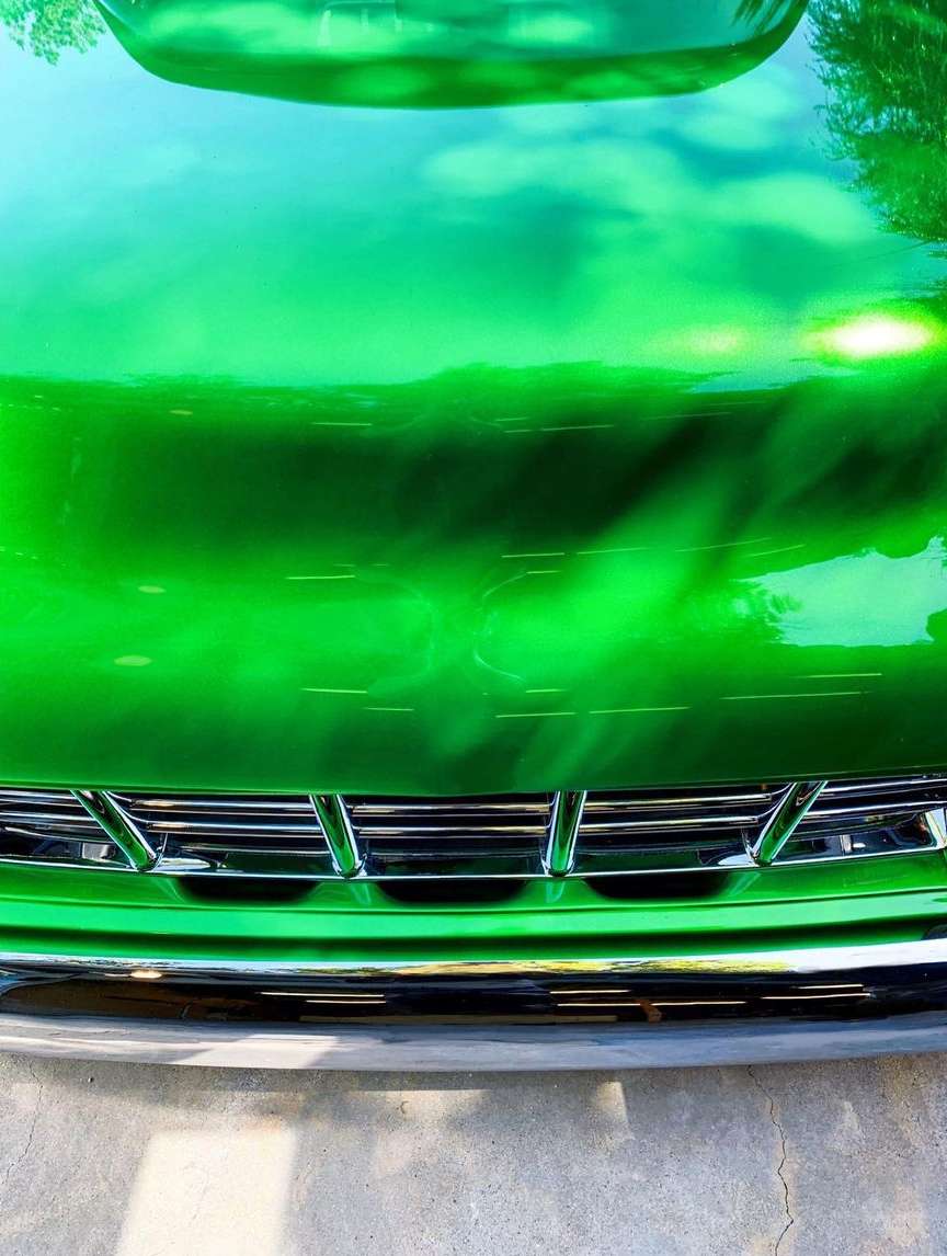 Close-up of a reflective green car hood with a chrome grill, capturing the sky's reflection and outdoor lighting.