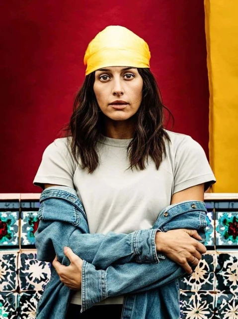 Portrait of a young woman with dark hair and a yellow headscarf, wearing a denim jacket, standing against a colorful tiled wall.