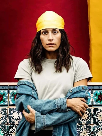 Portrait of a young woman with dark hair and a yellow headscarf, wearing a denim jacket, standing against a colorful tiled wall.
