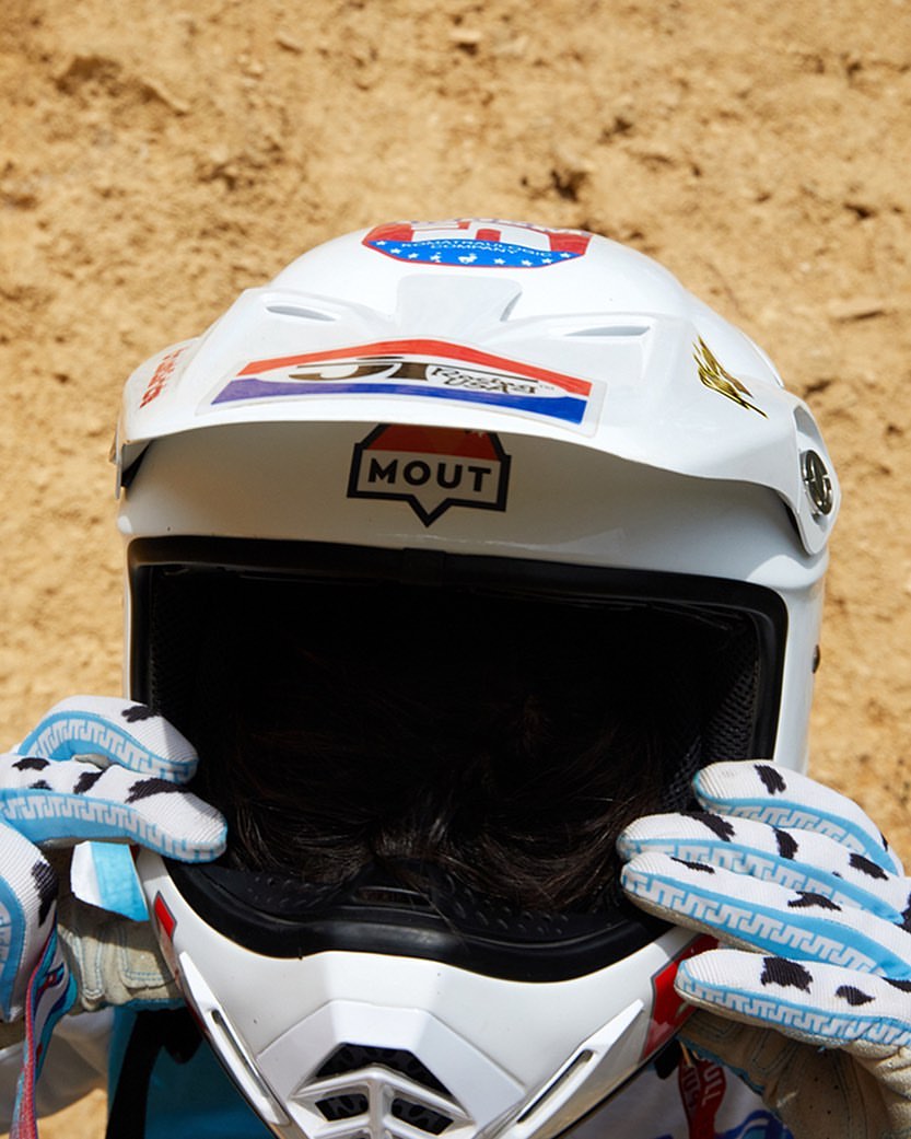 Close-up of a motocross racer's helmet and patterned gloves against a sandy backdrop, focusing on safety gear and sport details.