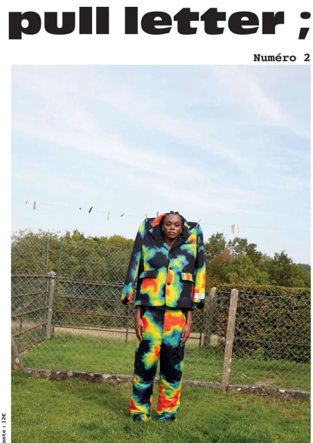 Editorial fashion shot of an adult female standing outdoors wearing a multicolored jacket and pants in a grassy yard setting.