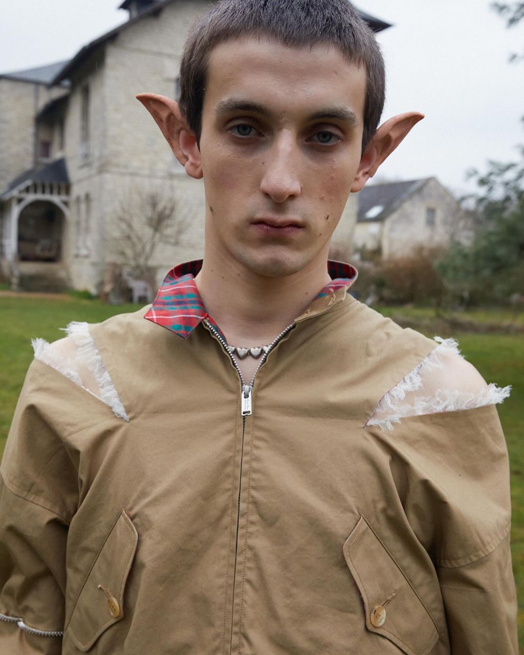 Portrait of a fair-skinned adult man with pointy ears wearing a beige jacket and silver necklace outside a light-colored building.