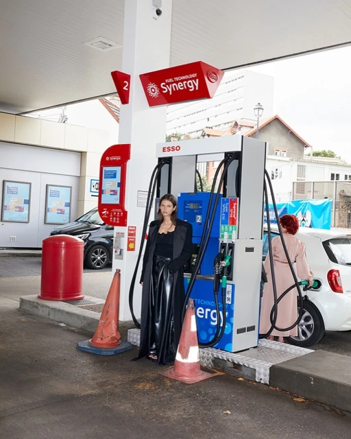 A woman in a black outfit poses at a gas station, another woman pumps gas into a white car. Traffic cones sit in the foreground.