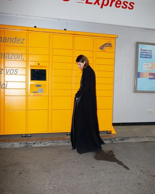 Adult woman in black coat walking past yellow automated delivery locker, outdoor daytime scene.