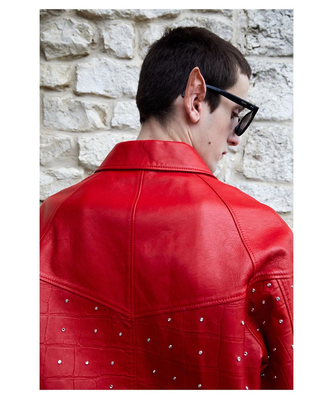 Man in red jacket with pointed ears and sunglasses stands against a stone wall looking away from the camera in a medium shot.