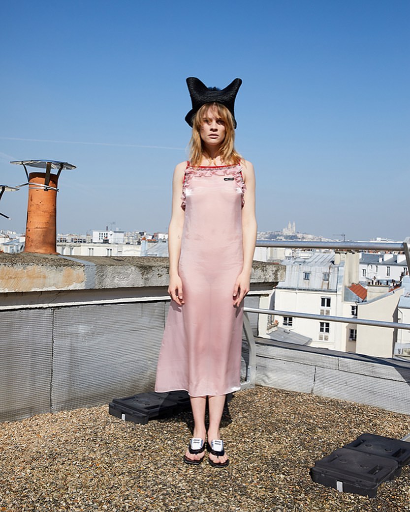 Model standing on a rooftop in Paris wearing a pink slip dress, black horned hat, and sandals, with a cityscape background.