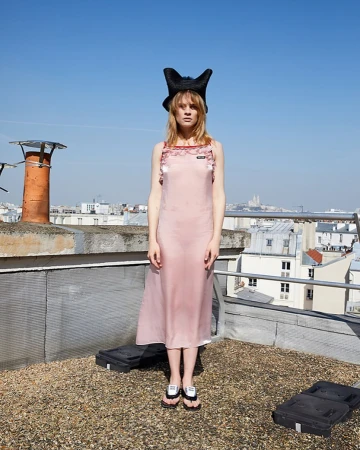 Model standing on a rooftop in Paris wearing a pink slip dress, black horned hat, and sandals, with a cityscape background.