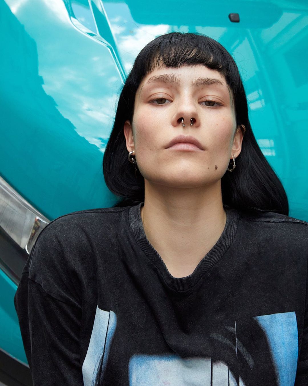 Close-up of a young adult female with dark hair and a nose ring, posing in front of a turquoise vintage vehicle on an overcast day.