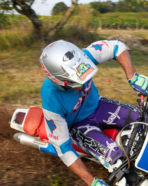 Motocross rider in full gear leans into a turn on a dirt track with green foliage and trees in the background, creating a dynamic scene.