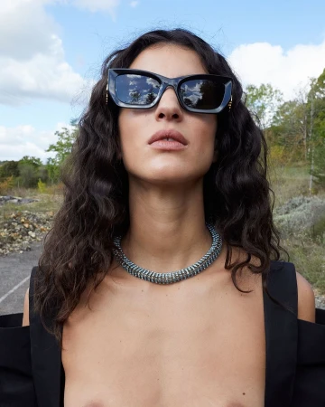 Close-up portrait of a woman wearing chunky black sunglasses and a textured silver necklace outdoors in Paris.