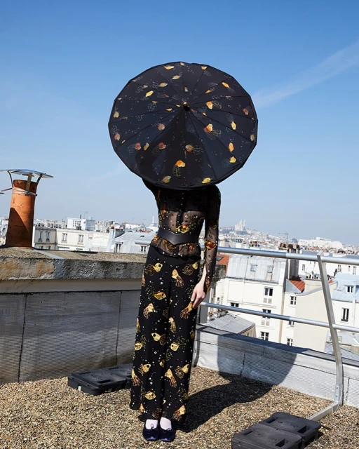 A woman stands on a rooftop in a sheer floral top, wide-leg pants and holds a printed black umbrella against a backdrop of Parisian buildings.