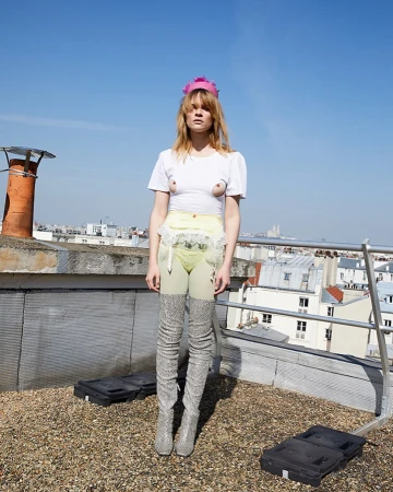 A slender woman with light hair stands on a rooftop in tall boots, a frilly skirt, and a small white t-shirt with buildings in the background.