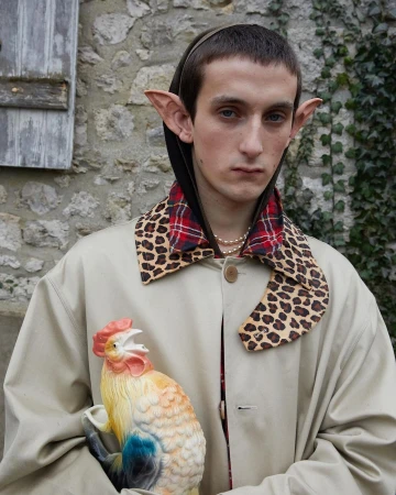 A young adult stands outdoors holding a rooster figurine, wearing elf ears and a unique coat with a patterned collar.