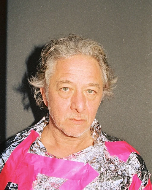 Close-up portrait of an adult male with gray hair, wearing shiny silver and pink foil-like garment, posing indoors against a gray wall.
