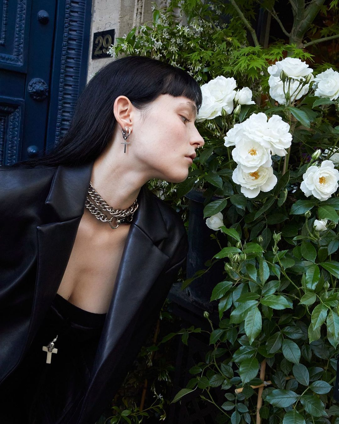 Adult female in black blazer stands near white roses with silver jewelry, including a chain necklace and cross earrings. 