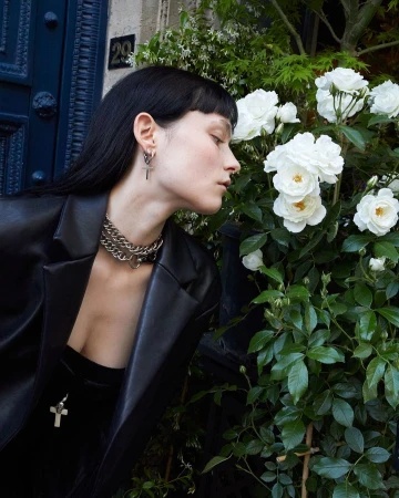 Adult female in black blazer stands near white roses with silver jewelry, including a chain necklace and cross earrings. 