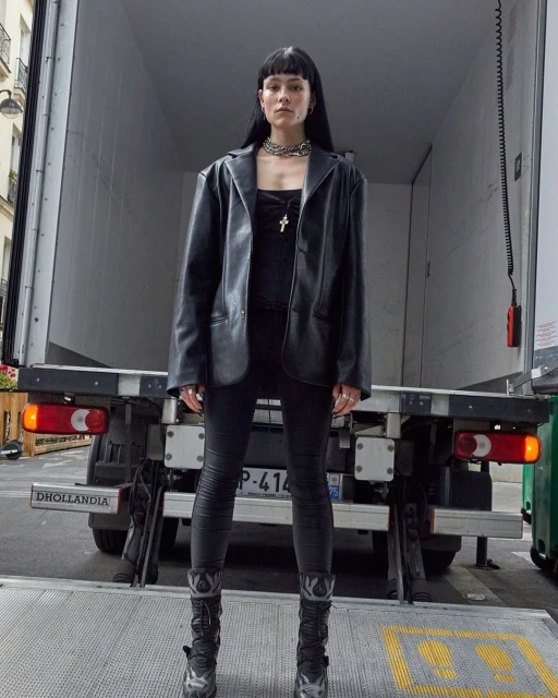 A young woman in a black leather jacket and leggings poses in front of a delivery truck; dark hair, neutral expression, urban setting.
