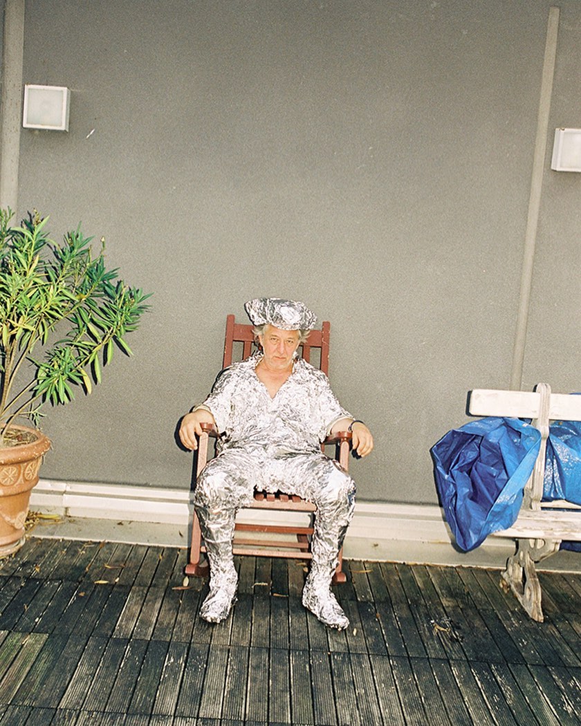 An older adult sits on a wooden rocking chair wearing a full outfit made from shiny silver material and a matching hat, outdoors.