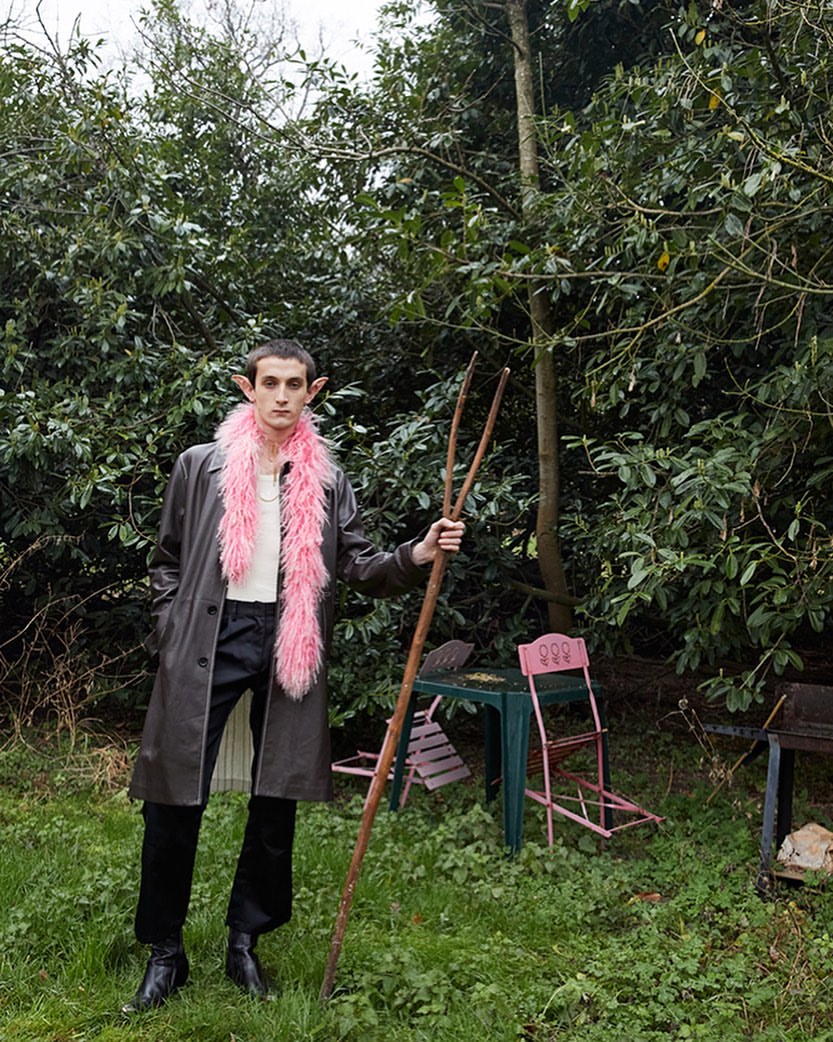 Young man in a black coat and pink faux fur stole posing in a garden with a pitchfork, leaning against a backdrop of lush greenery.