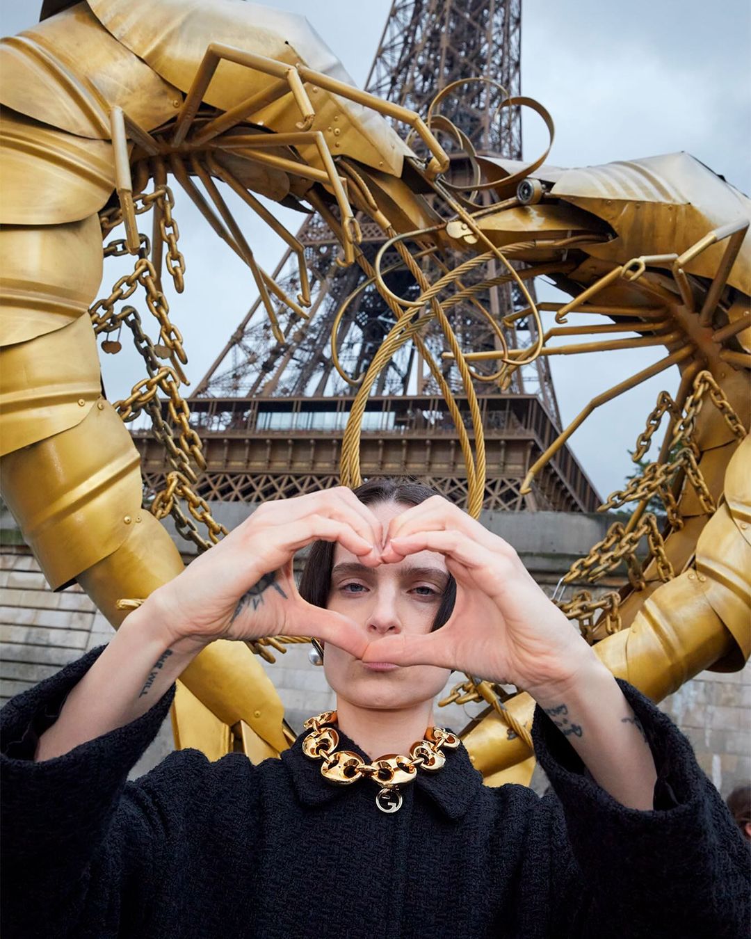 Woman makes heart shape with hands in front of Eiffel Tower and a golden shrimp sculpture wearing black jacket and gold necklace.