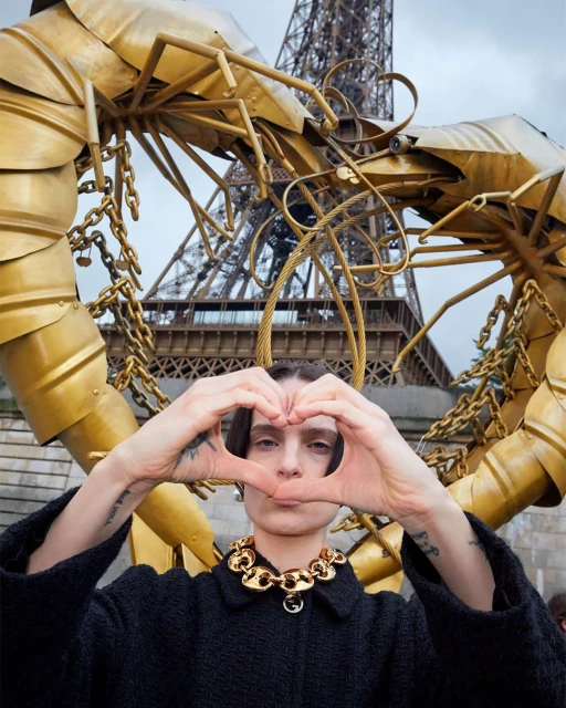 Woman makes heart shape with hands in front of Eiffel Tower and a golden shrimp sculpture wearing black jacket and gold necklace.