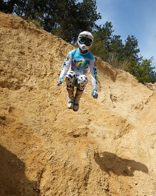 Person in full protective gear jumping over a sand dune on a sunny day with green trees and a clear blue sky in the background.