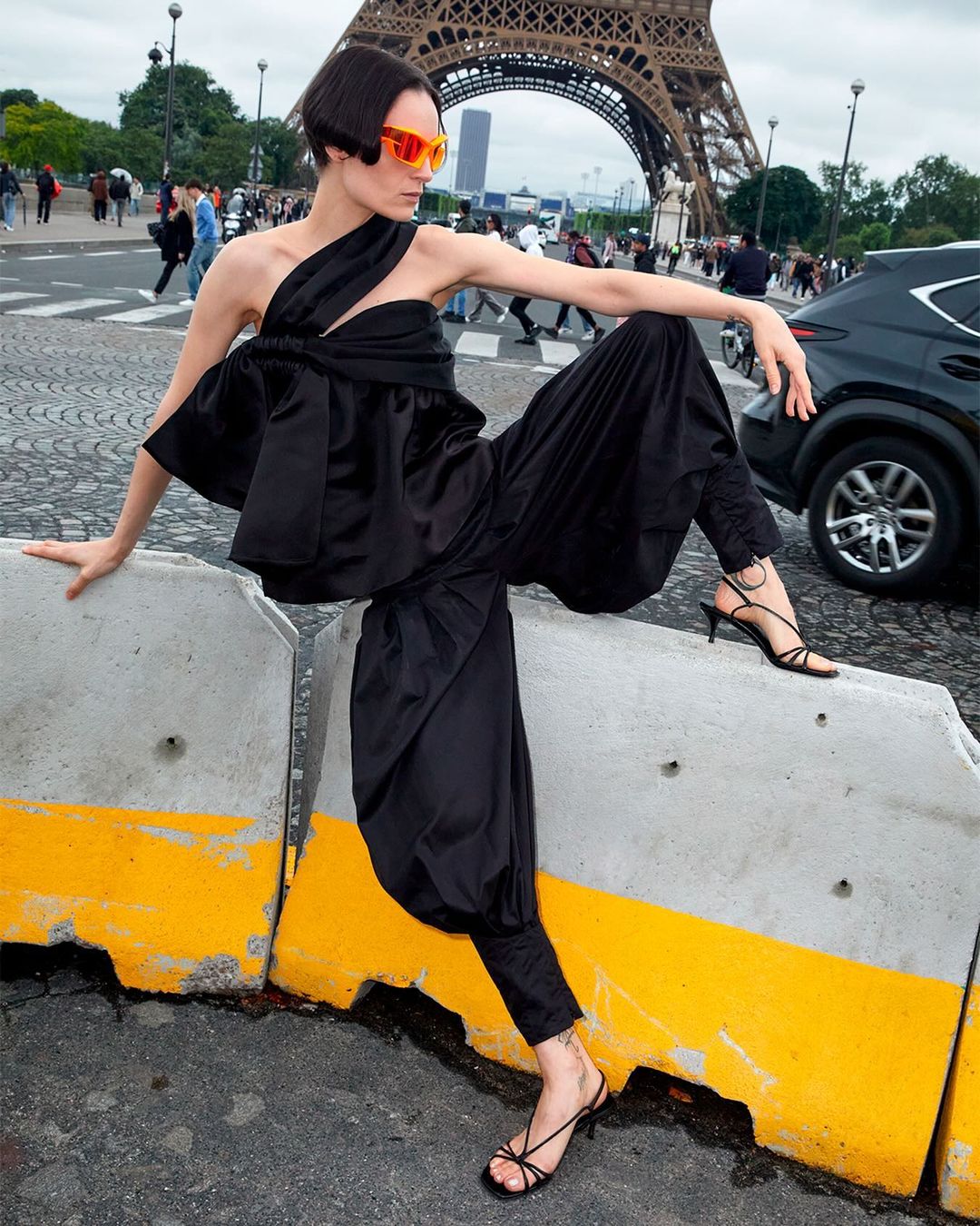 A woman in an avant-garde black outfit poses on a concrete barrier in front of the Eiffel Tower in Paris, wearing orange sunglasses.