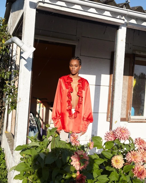 A stylish young Black woman poses in a red ruffled tunic top amidst vibrant dahlia flowers outside a white-painted, rustic house on a sunny day.