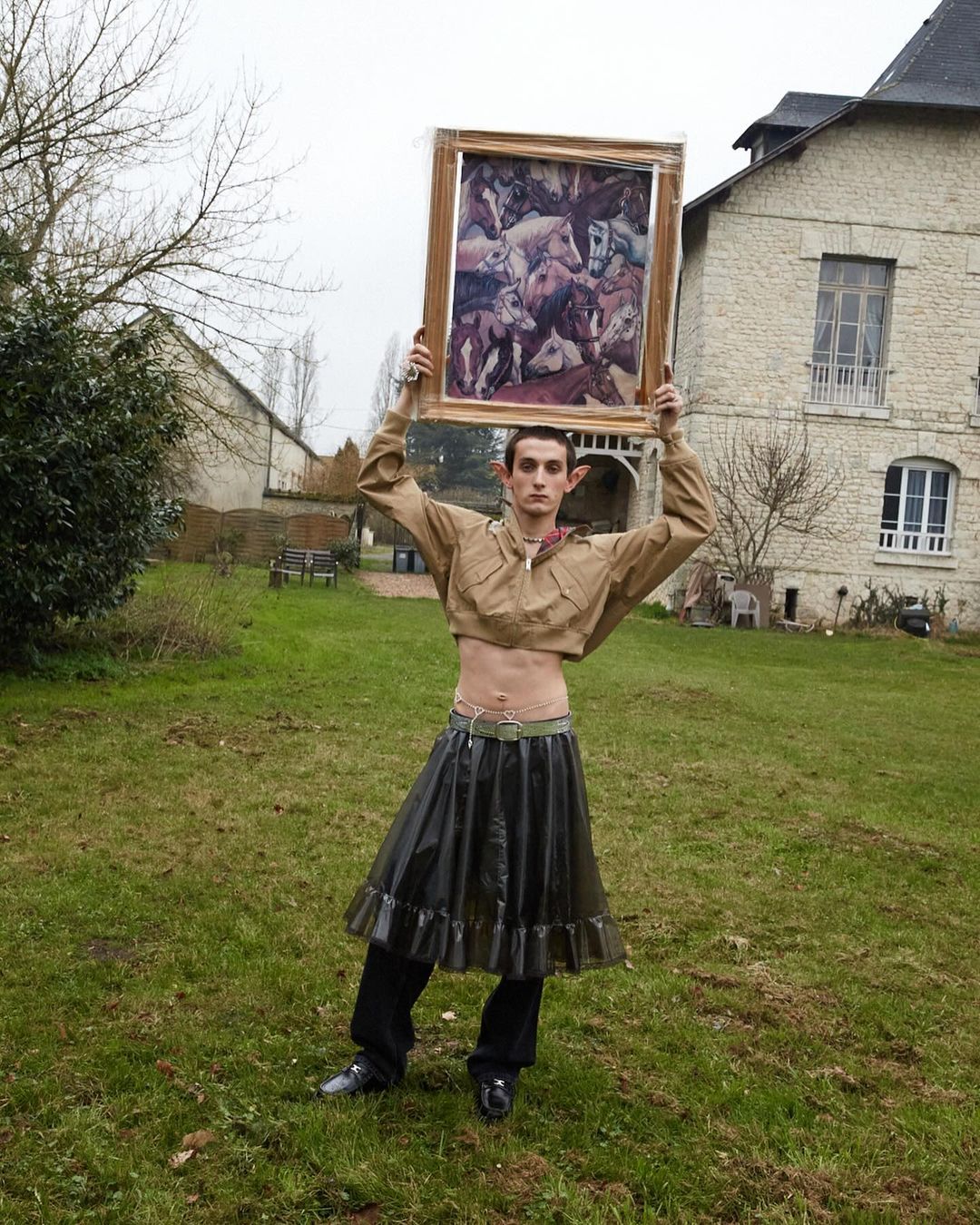 An adult posing outdoors holding a framed print of a horse painting, wearing a crop top and clear plastic skirt in a yard setting.