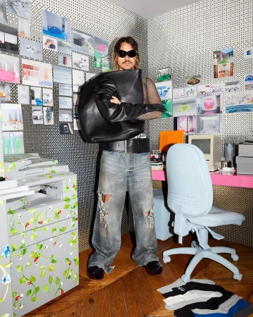 An adult male poses in an office with a black leather bag, ripped jeans, and sunglasses, surrounded by a computer, chair, and patterned wallpaper.