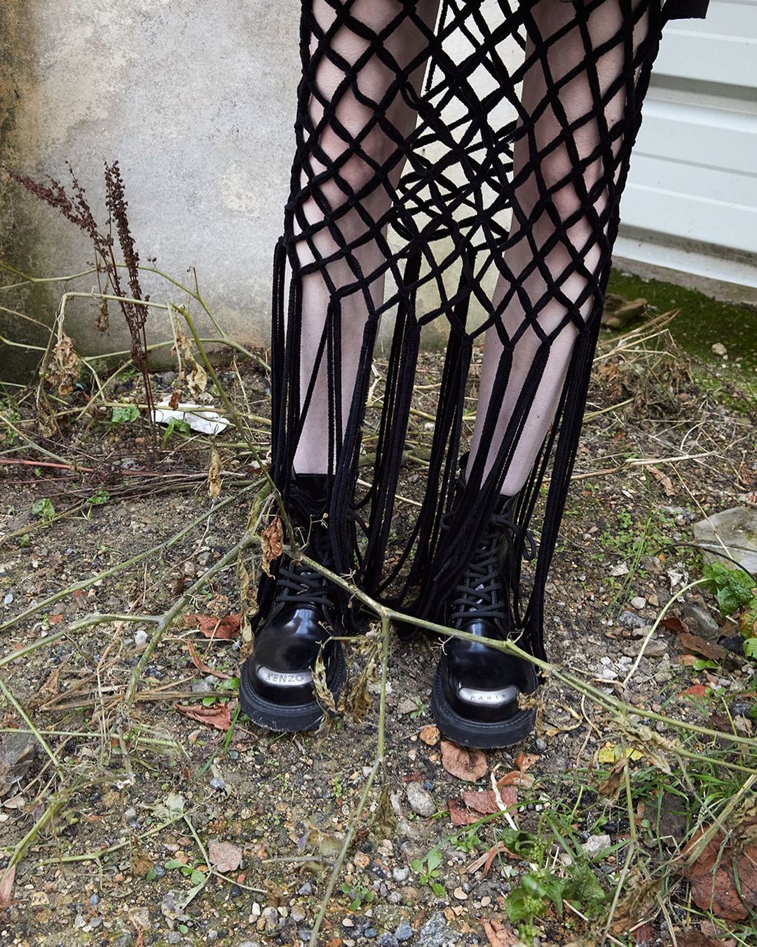 Fashionable black lace-up boots and net-like fringed garment worn outdoors, standing on dry foliage near a weathered concrete wall.