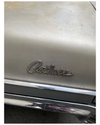 Close-up of a weathered vintage car hood featuring the metallic script emblem Cutlass and chrome bumper trim.