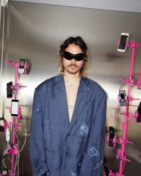 Man with dark curly hair and sunglasses wearing an oversized blue patterned blazer against a metallic background
