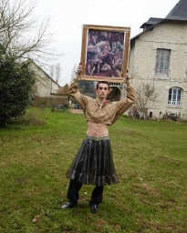 A man wearing a cropped jacket and dark skirt holding up a framed horse painting in a grassy yard.