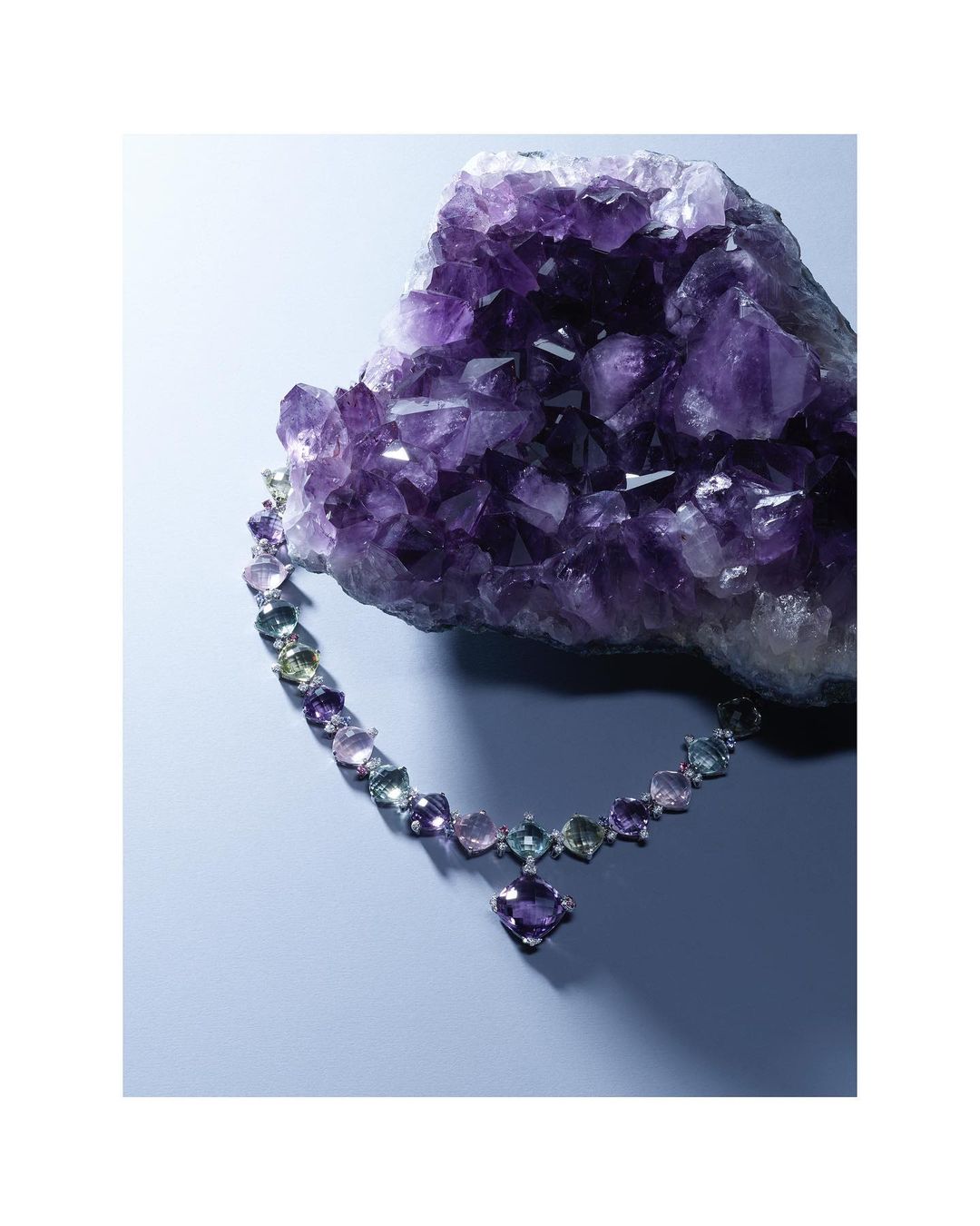 Elegant necklace with square-cut gems, set against a sparkling purple amethyst geode, on a light blue surface with soft studio lighting.