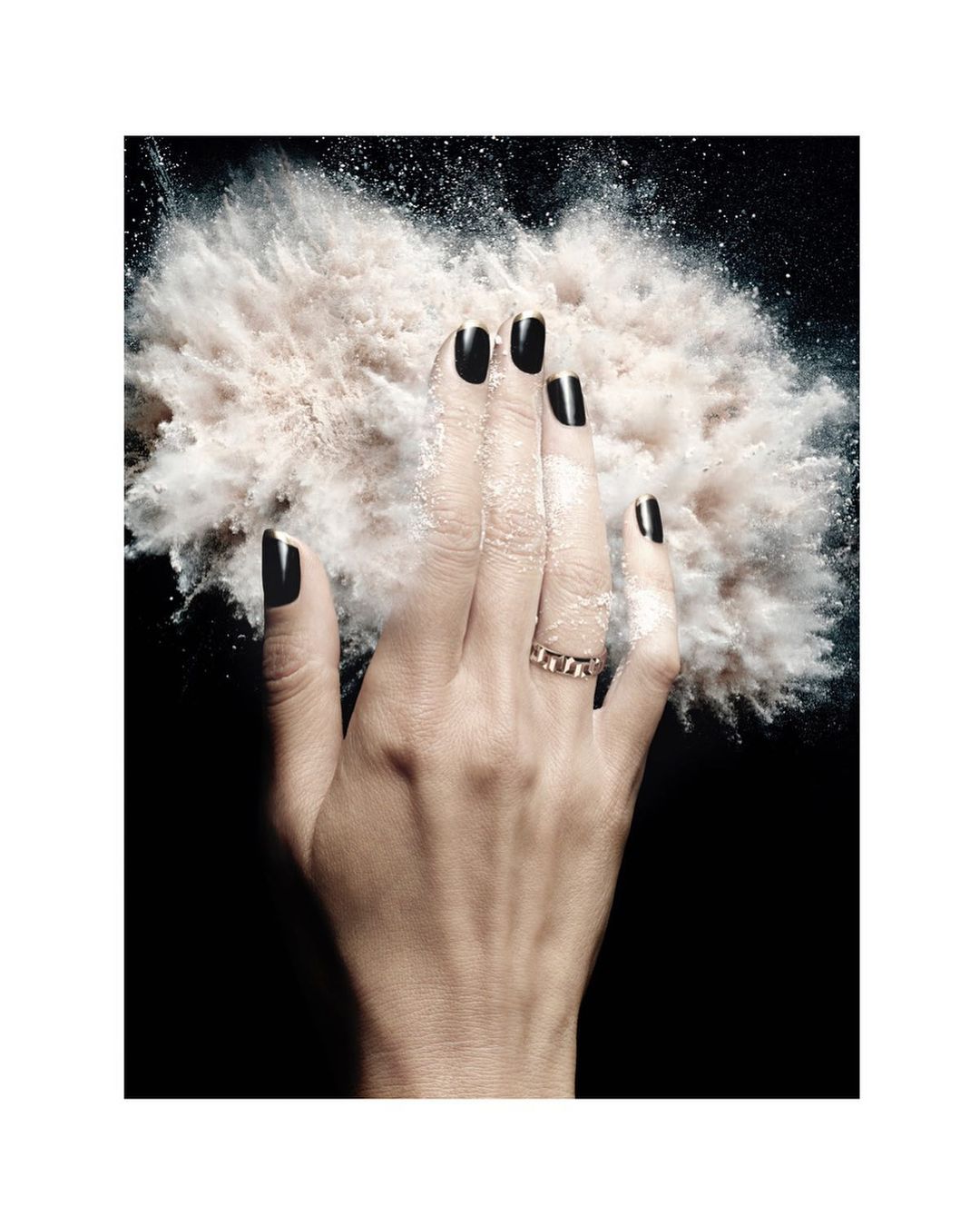 Close-up of a hand with black nail polish and a gold ring surrounded by a cloud of white powder against a black background.