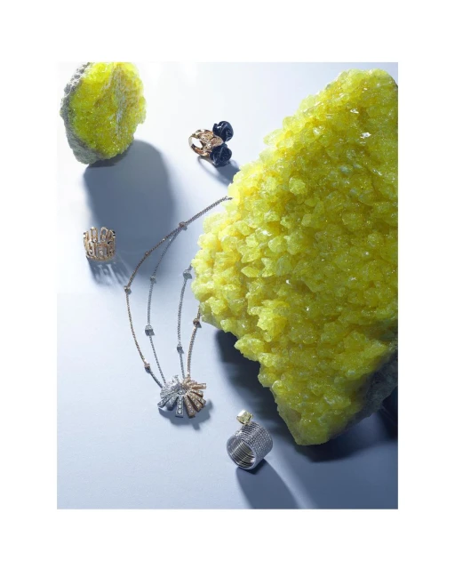 Fine jewelry including a necklace and rings is displayed on a white surface with a light blue gradient, accented by a yellow mineral.