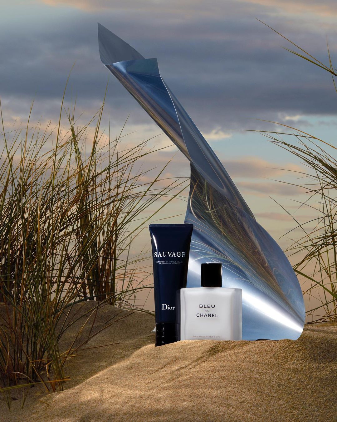 Dior Sauvage and Bleu de Chanel bottles on sand with an abstract metal sculpture and grassy dunes against a cloudy sky backdrop.