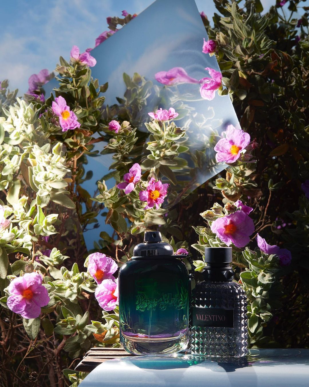 Two perfume bottles are placed among blooming pink rockrose flowers with a mirror reflecting the blue sky and white clouds in a natural setting.