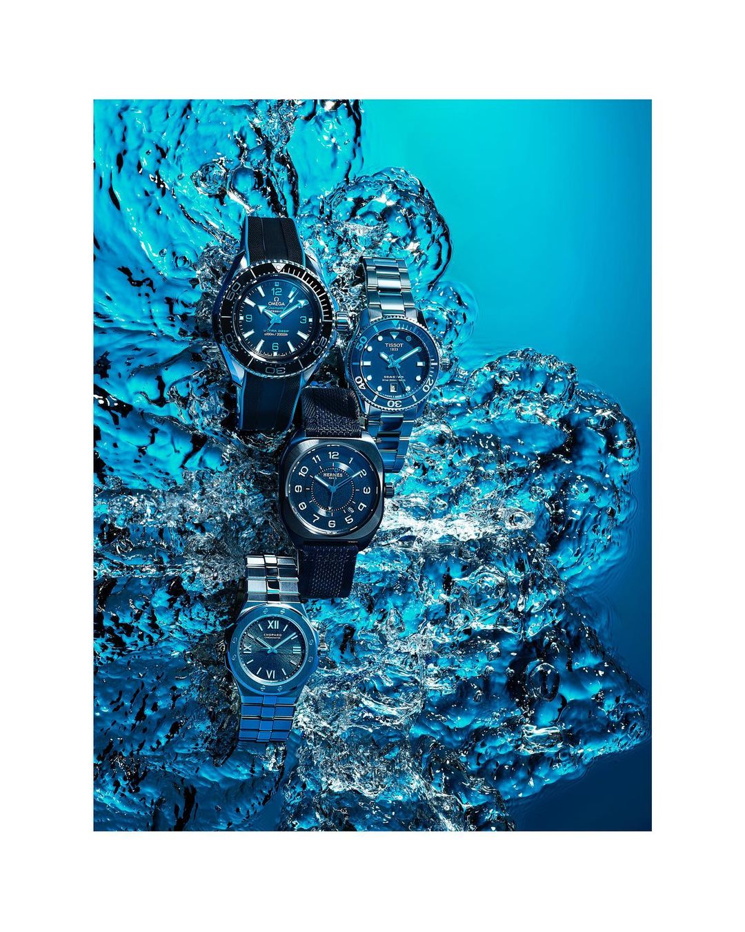 Four luxury wristwatches are displayed in a still life arrangement surrounded by splashing blue water, creating a dynamic and visually appealing composition.
