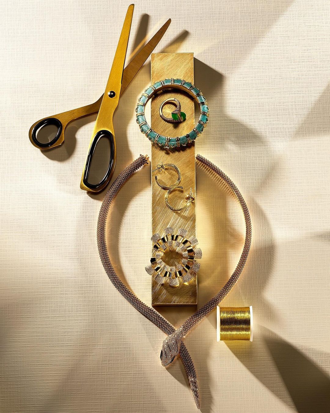 Overhead studio shot of gold scissors, rings, gold earrings, snake necklace, and gold thread on a gold jewelry stand.