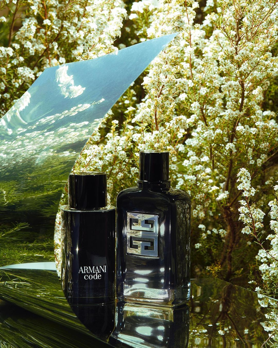 Two perfume bottles, Armani Code and Givenchy, are staged among blooming white flowers and reflective surfaces in an outdoor setting.