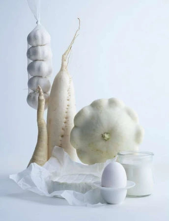 Still life shot of white vegetables: garlic, radish, pattypan squash, egg, and cheese against a white background creating a minimalist aesthetic.