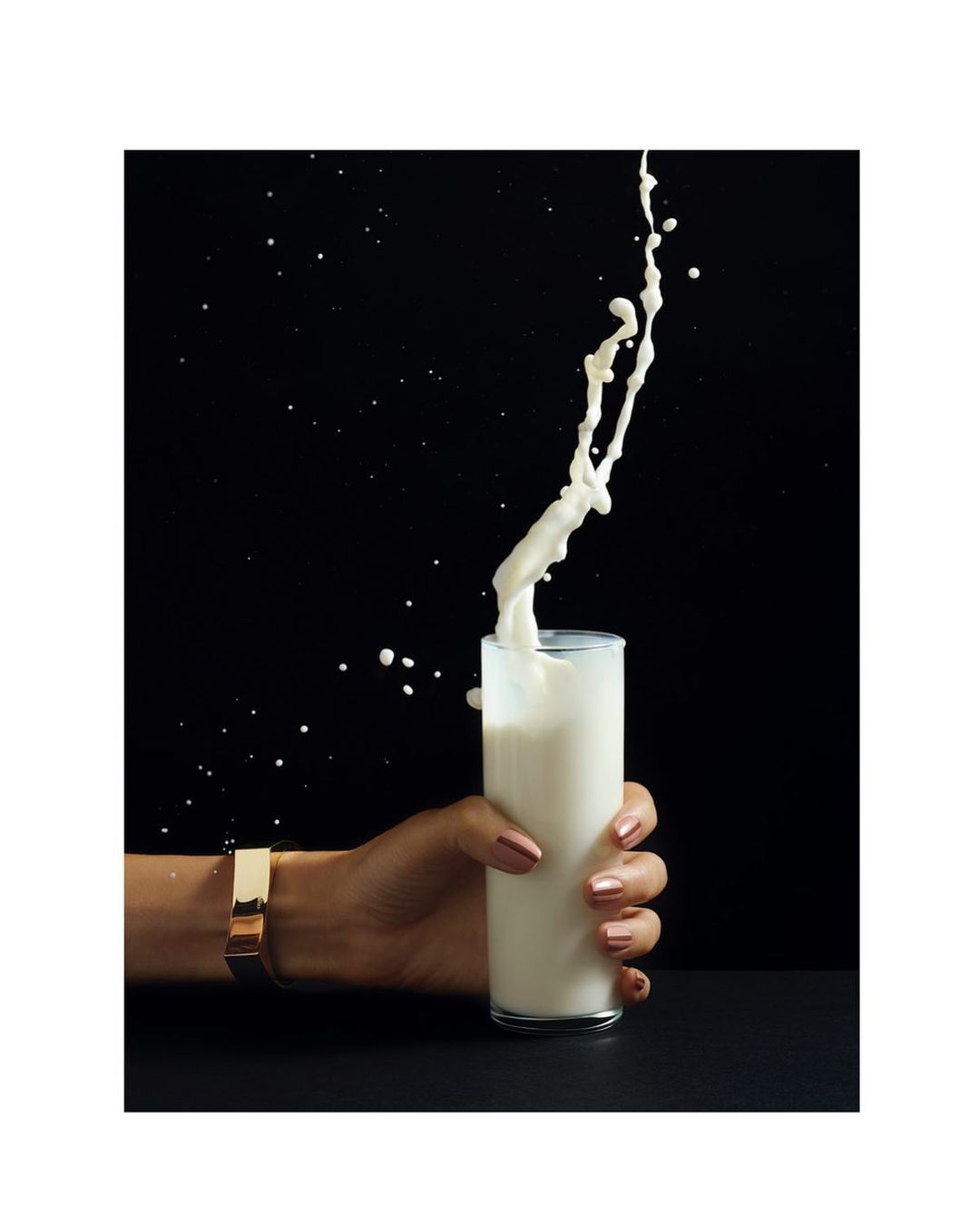 A woman's hand wearing a gold bracelet holds a glass as milk is poured in and splashes against a black background.