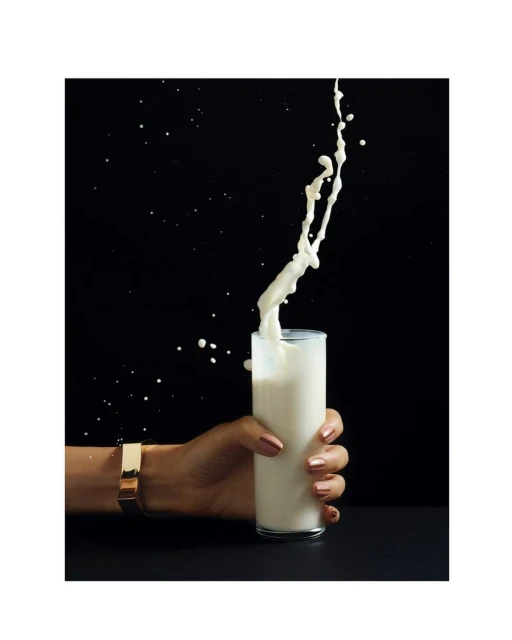 A woman's hand wearing a gold bracelet holds a glass as milk is poured in and splashes against a black background.