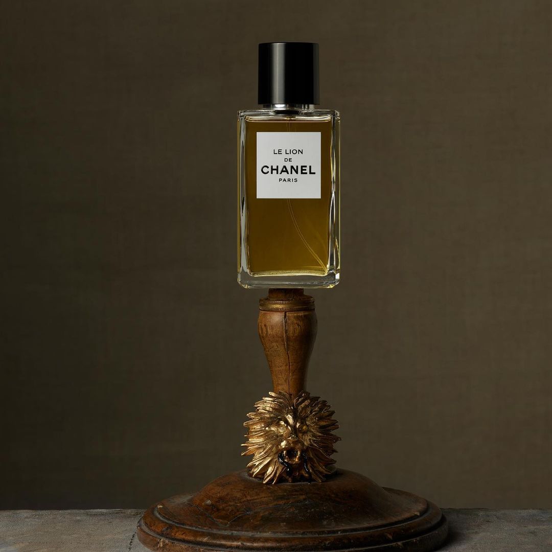 Perfume bottle of Le Lion de Chanel Paris on a wooden pedestal with a golden lion head against a muted brown background.