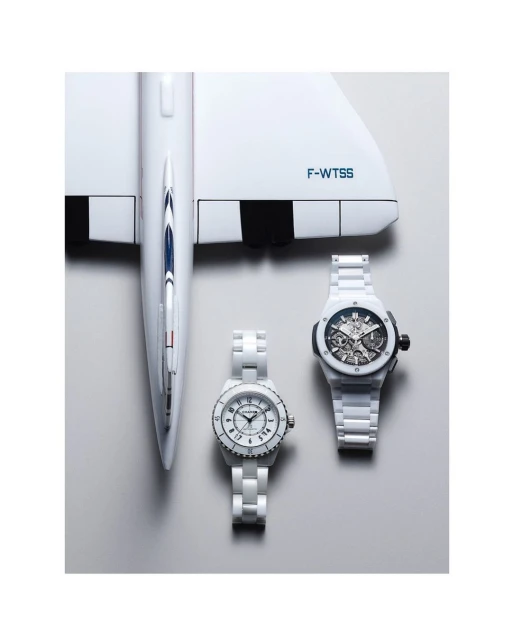 Elevated studio shot of two white ceramic wristwatches on a gray surface next to a white model airplane with black tail markings.