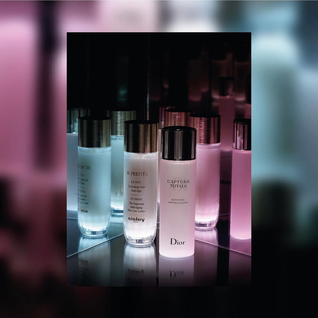Cosmetic bottles from Dior and Sisley are displayed on a mirrored shelf in a dark environment with soft pink and blue lighting.