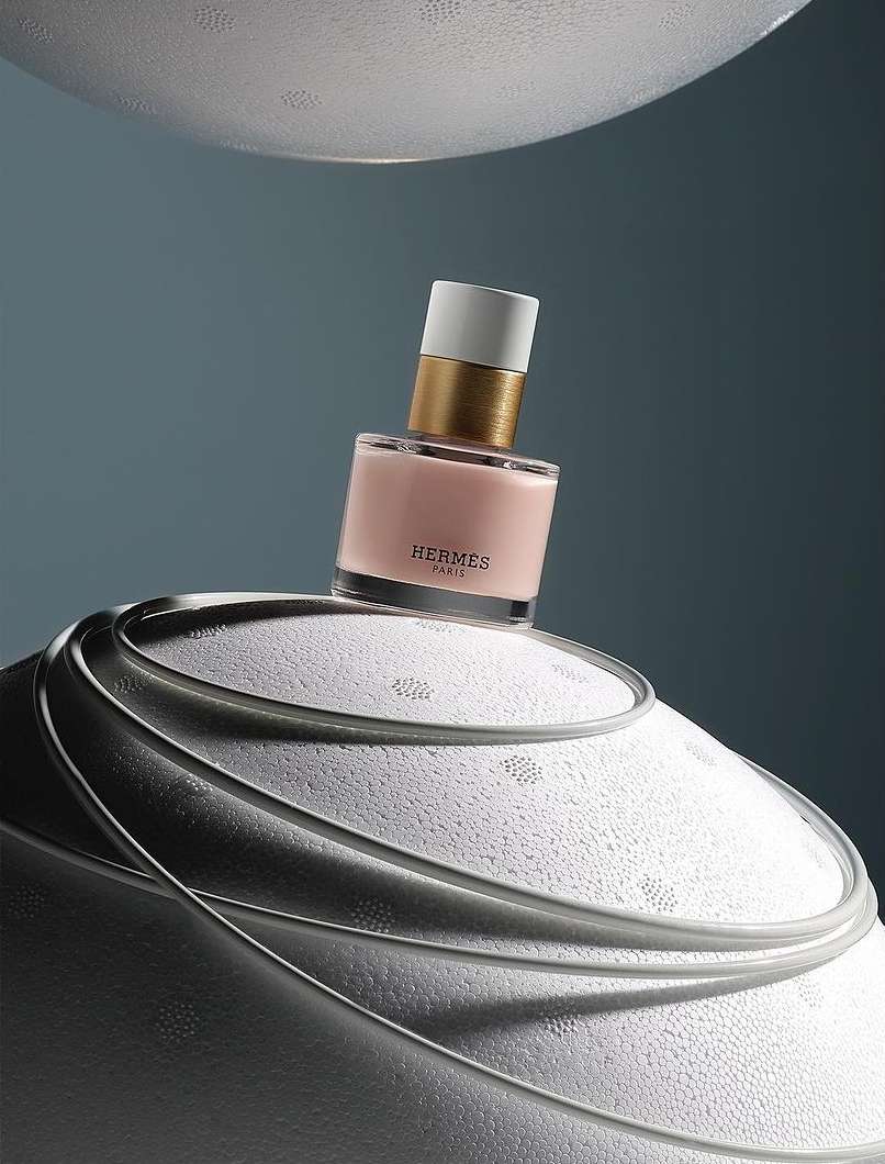 Hermes perfume bottle on a textured white surface against a blue backdrop. The bottle features a white cap, gold neck, and light pink liquid.