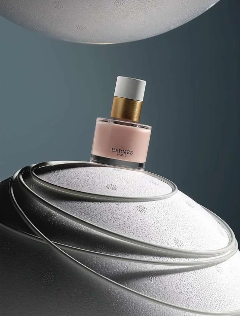 Hermes perfume bottle on a textured white surface against a blue backdrop. The bottle features a white cap, gold neck, and light pink liquid.